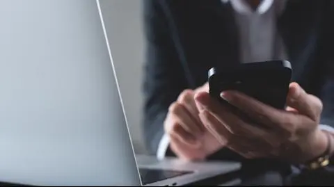 shutterstock_2514190811 Close up, bizneswoman korzystająca ze smartfona podczas pracy na laptopie w biurze. Bizneswoman w biznesie siedząca przy stole za pomocą smartfona, surfująca po Internecie