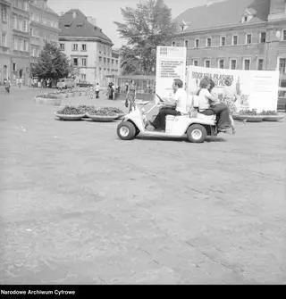 
Turyści jadący elektrycznym pojazdem typu "Melex". W tle plansza "Dom Mody Astra - Warszawa ul. Pankiewicza 4 (1974 r.)