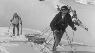 Dwaj ratownicy na nartach, jeden z nich ciągnie sanie do transportu rannego. W tle budynki na Kasprowym Wierchu. Zdjęcie wykonane przez propagandę niemiecką, 1942. 
(http://www.audiovis.nac.gov.pl/obraz/21753/e59e623c7b84d21f9b2fa949c453f764/)
